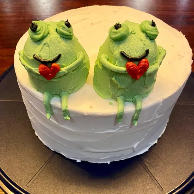 White cake topped with two buttercream frogs holding hearts